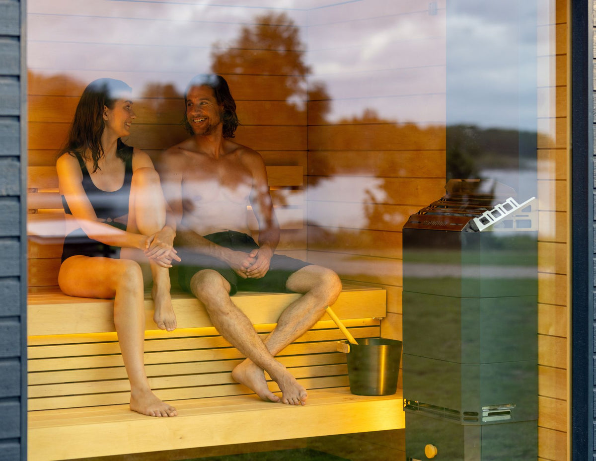 Couple on the bench of a glass-front sauna with the Saunum Air 5 heater beside them