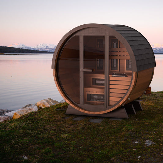 Finnmark FD-6 cedar barrel sauna standing on a shoreline at dusk with mountains behind