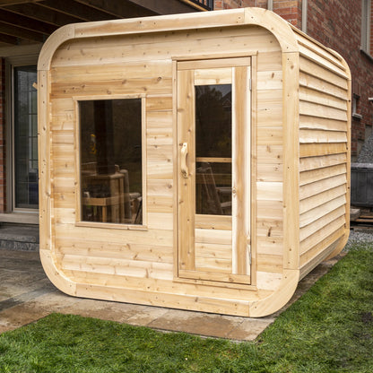 CT Luna cedar sauna on a lawn beside a brick house, three quarter view showing the door and window wall
