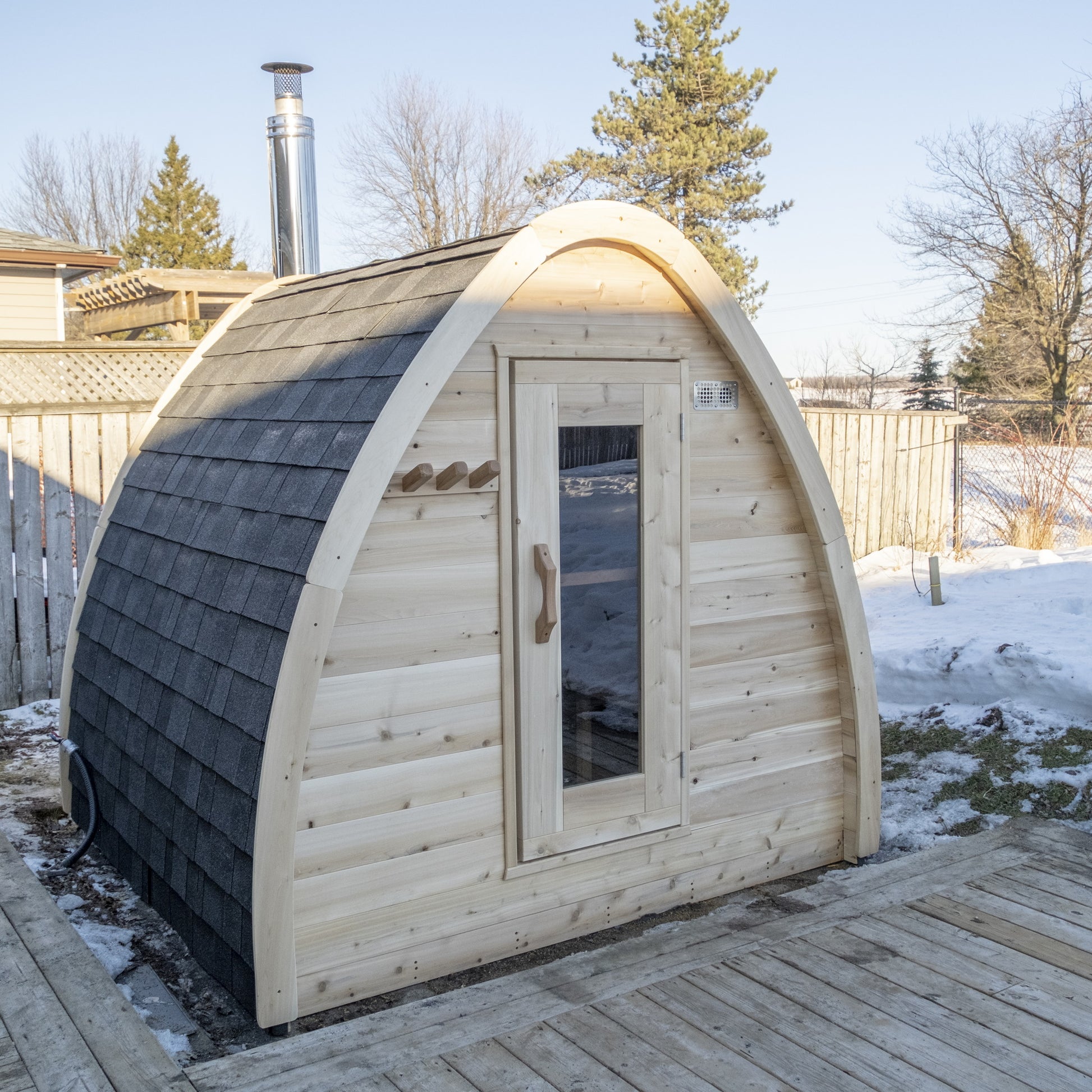 Canadian Timber CT MiniPOD outdoor pod sauna in a snowy backyard, three-quarter view of the raindrop shape, black shingle roof and chimney