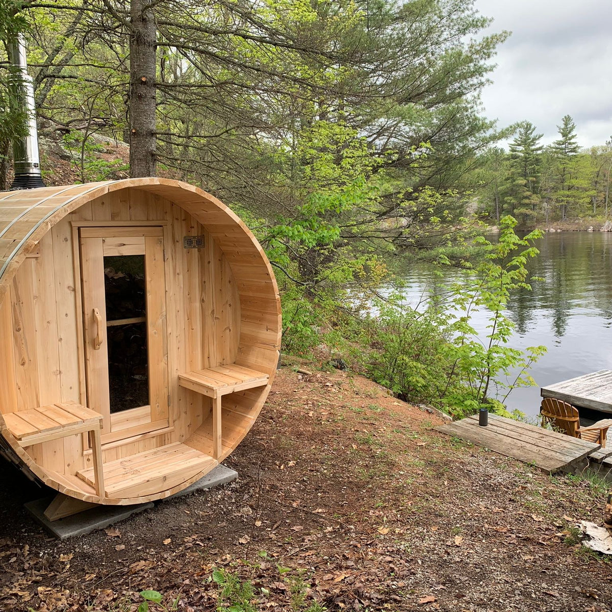 CT Serenity barrel sauna on a wooded lakeside slope, porch and outside benches facing the water