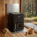 Narvi Kaamos 20 black wood-burning sauna stove with heat shield beside a forest window, logs and bucket alongside