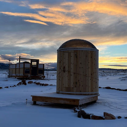 The Skylight barrel washroom on a timber platform in deep snow at sunset, its dome on top and an outbuilding on the plain behind.