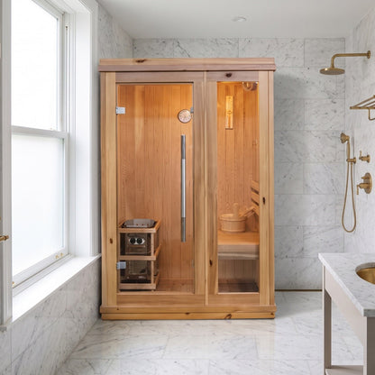 SunRay Aston indoor sauna for 1 to 2 in cedar, glass door with a long steel pull and the Harvia heater inside, against white marble walls next to a tall sash window