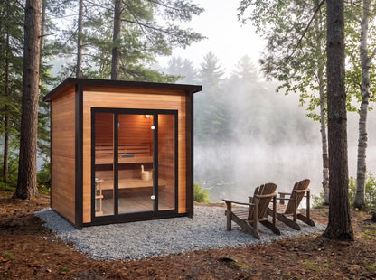 SunRay Cayman 4-person outdoor sauna on a gravel pad among pines by a misty lake, two Adirondack chairs beside the black-framed glass front