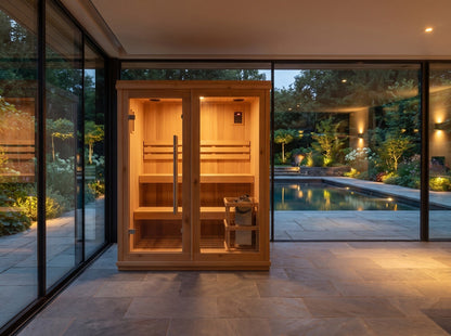 SunRay Charleston 4-person cedar indoor sauna in a glass-walled pool house at dusk, two-tier benches and heater lit behind the glass front