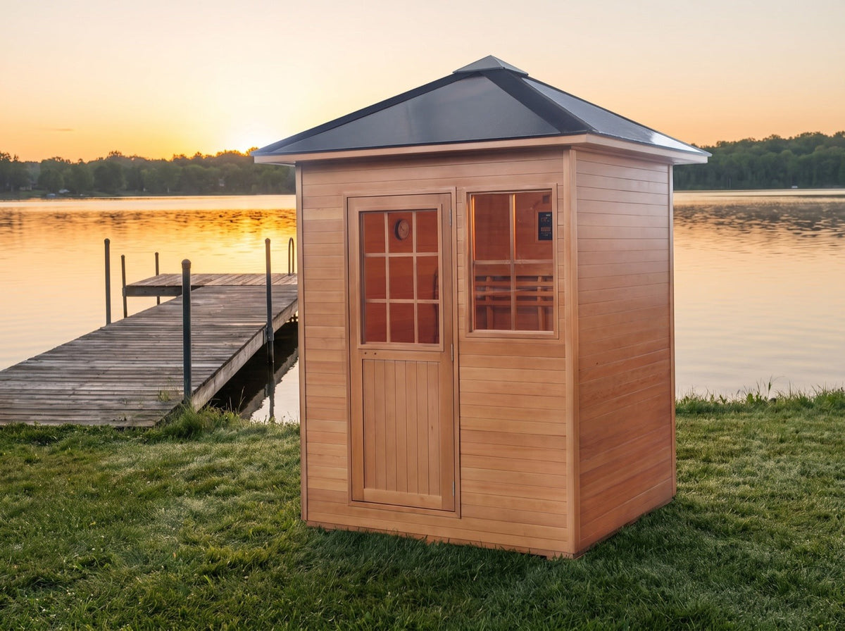 SunRay Freeport outdoor sauna on a lawn by a lake at sunset, with its black pitched roof, wooden door with a multi-pane window and front window