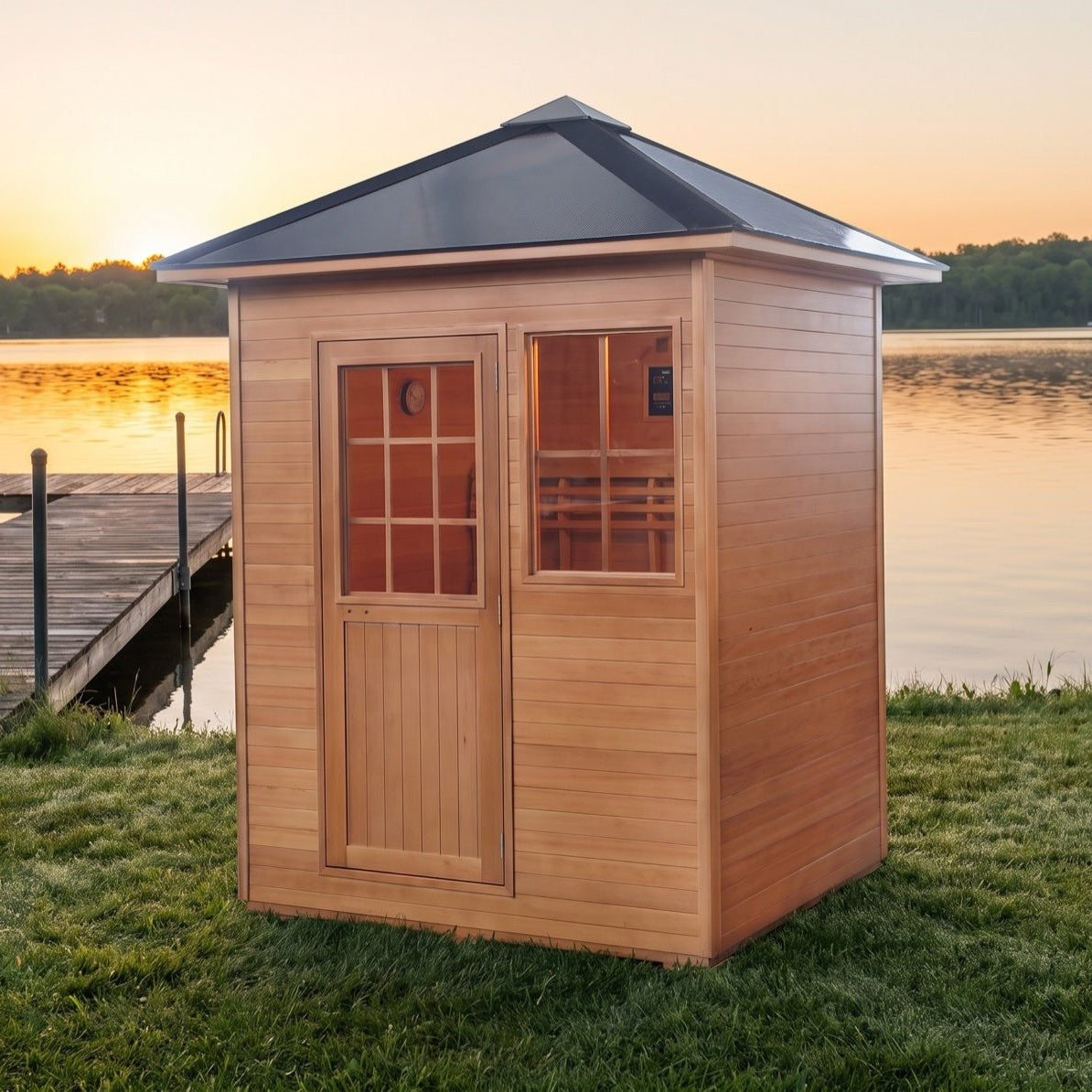 SunRay Freeport outdoor sauna for 3 in hemlock, wooden door with a multi pane window beside the front window under the black pitched roof, on a lakeside lawn next to a dock at sunset