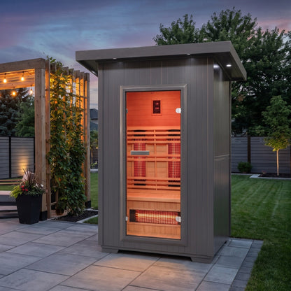SunRay Logan outdoor infrared sauna for 2 with a charcoal exterior and hemlock interior, glass door glowing with the heater panels, on a stone patio beside a lit pergola at dusk