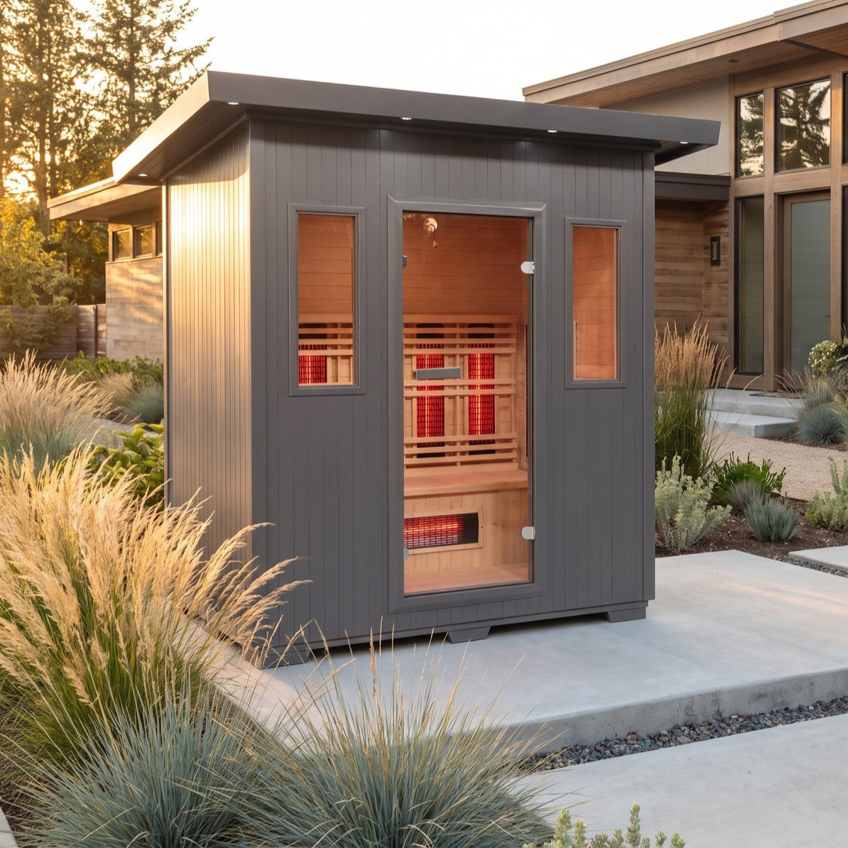 SunRay Pacific outdoor infrared sauna for 4 with a charcoal exterior and hemlock interior, glass door between two windows, beside a modern house among ornamental grasses