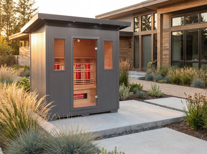 SunRay Pacific outdoor infrared sauna on a concrete path beside a modern house, among ornamental grasses