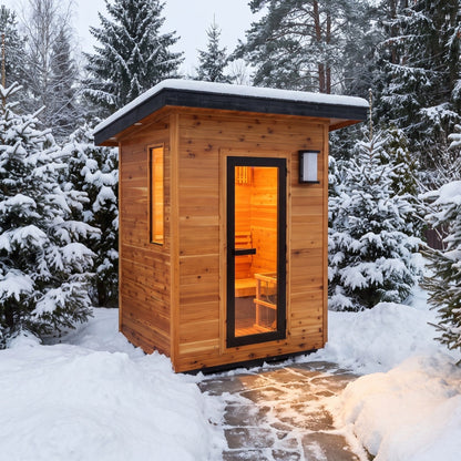 SunRay Seneca outdoor sauna for 3 in western red cedar, black framed glass door, side window and wall lantern under a snow topped roof, on a cleared stone path among snowy evergreens