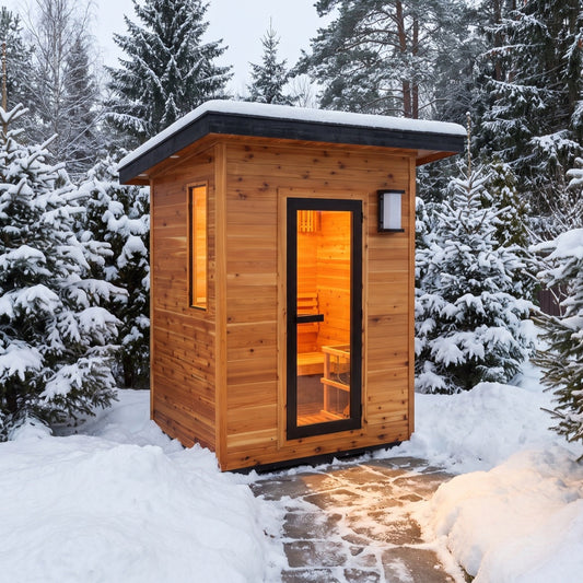 SunRay Seneca outdoor sauna for 3 in western red cedar, black framed glass door, side window and wall lantern under a snow topped roof, on a cleared stone path among snowy evergreens