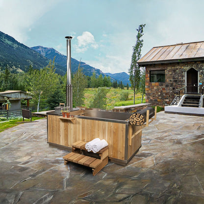 The Starlight Wood Burning Hot Tub on a flagstone patio below mountains, chimney at the stove end, shown with the optional aluminum interior and roll-up cover.